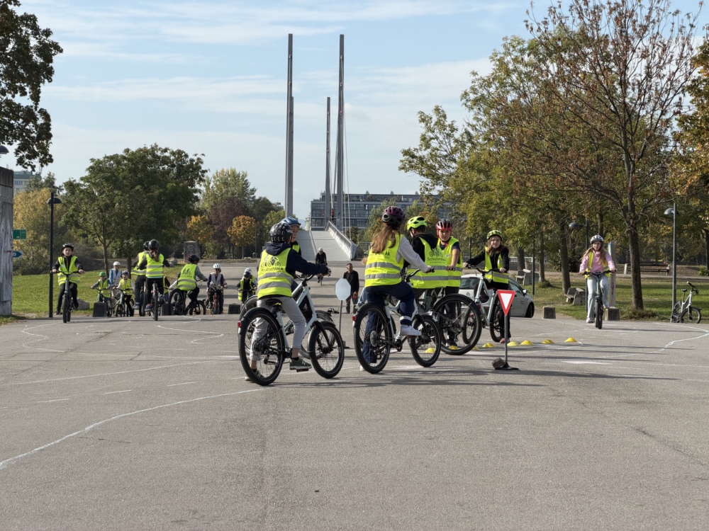 Kinder mit Warnwesten üben Radfahren in einer Übungszone mit Hindernissen und Markierungen.